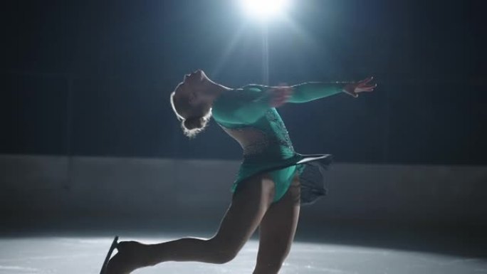 晚上，穿着裙子的美丽女孩溜冰者在聚光灯下独自在溜冰场上滑冰。运动员进行了一次后退旋转。