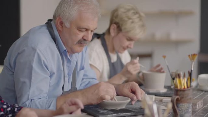 一群在陶器大师班上的老人一起在陶杯上雕刻和切割绘画，以制造陶瓷餐具
