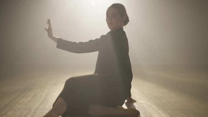 热情苗条优雅的芭蕾舞演员在背光雾中坐在地板上跳舞。白人年轻女子在舞台上聚光灯下表演现代国标舞的镜头。