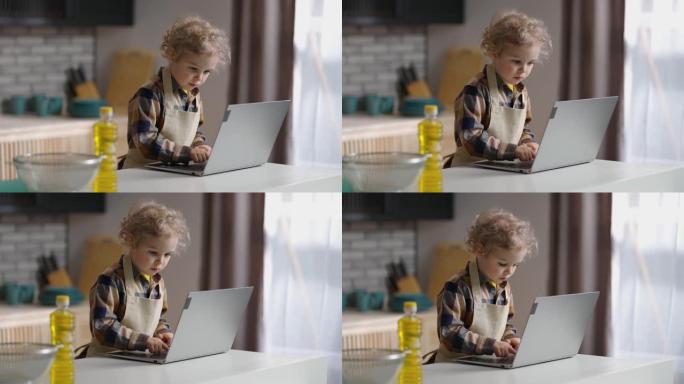 蹒跚学步的厨师穿着围裙正在用笔记本电脑在互联网上寻找食谱，小男孩在家庭厨房