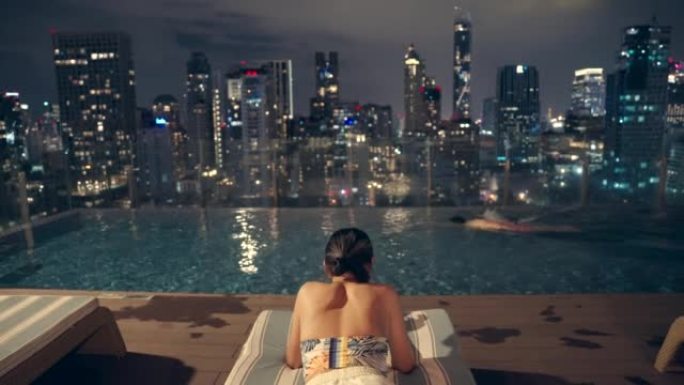 晚上，一名亚洲男子游泳和女子拿着手机坐在一栋高层建筑的游泳池里，俯瞰着这座城市。