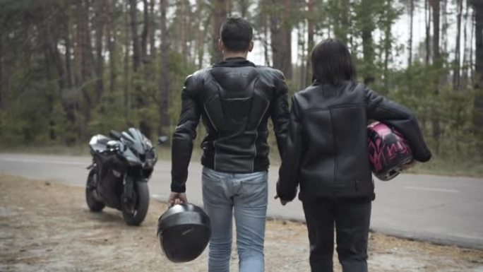 自信的几个摩托车手牵着手沿着路边走过黑色时尚摩托车的后视图。镜头跟随跨种族的爱男人和女人在户外漫步。