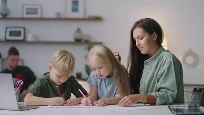 一位有两个孩子的母亲坐在客厅的桌子旁画画。家庭用铅笔画画