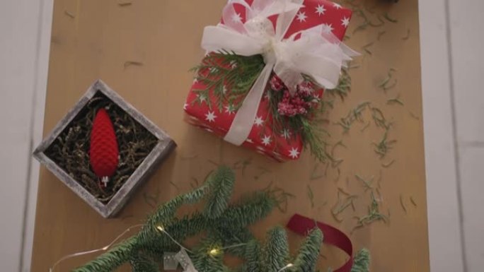 俯视图平面图。完全可见带有装饰品的桌子。女性双手将用手纸包裹的圣诞礼物放在木桌上并完成。包扎胶带和绑