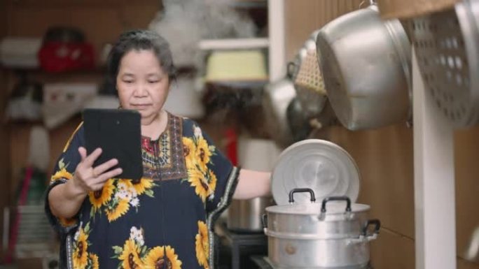 亚洲高级女性在家准备食物并检查平板电脑上的食谱或视频教程