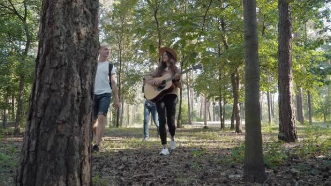 美丽的女人在夏天的森林里和朋友一起散步时弹吉他的极宽镜头。积极的白种人游客离开，英俊的男人通过双筒望