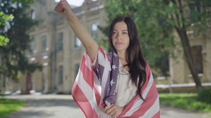 在大学校园里，一个自信的年轻女子在美国国旗上做着自由的手势。严肃美丽的黑发学生摆姿势自由在大学的背景