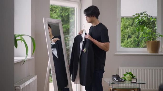 年轻的新郎在客厅里照镜子试穿婚纱。亚洲男子欣赏倒影衣架的侧视图，饰有优雅的胸衣。准备结婚。