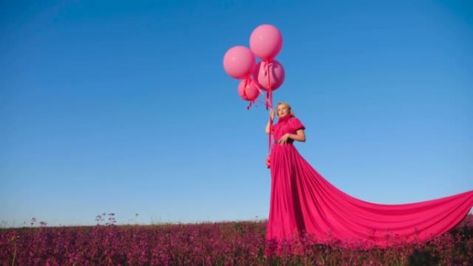 穿着粉红色长裙的时尚美女站在田野上，夏天在蓝天背景下，粉红色的花朵和氦气球站在田野上