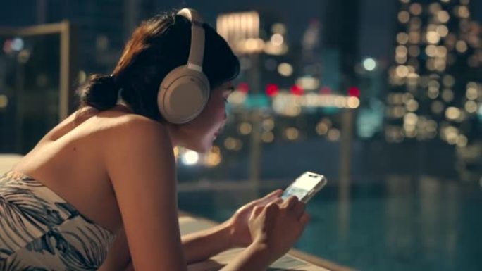 她听音乐，用手机玩网上购物。泰国曼谷城景高楼上的游泳池区。