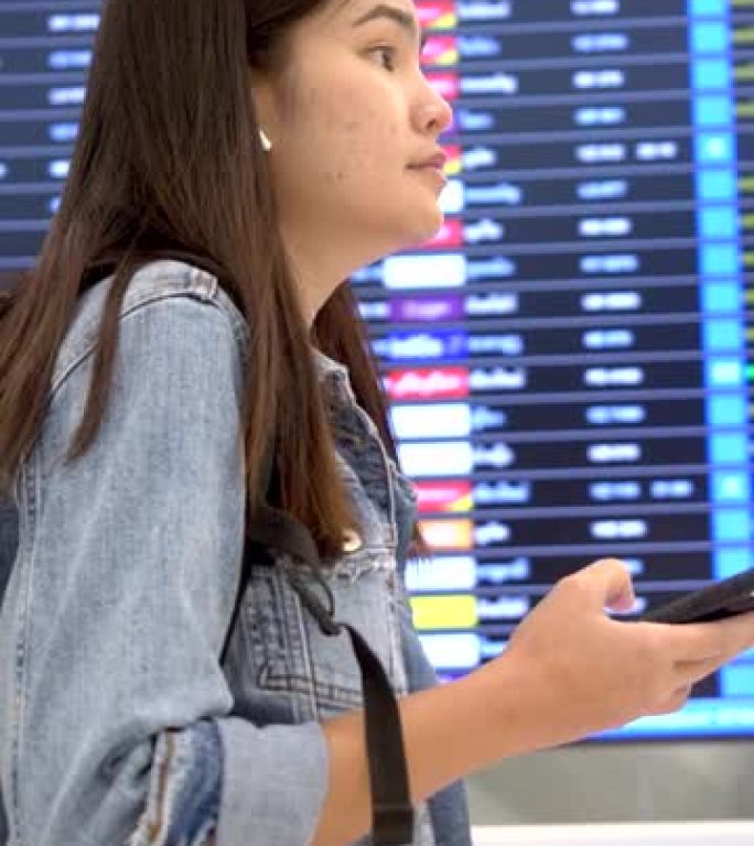 垂直格式视频: 穿着蓝色牛仔裤的亚裔女乘客在乘客大楼内拖着带轮行李箱