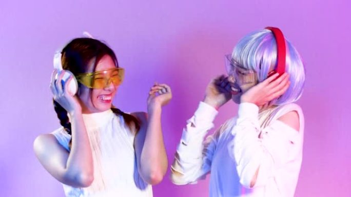两个穿着白衬衫的年轻亚洲女性戴着耳机听音乐并在紫色屏幕背景上摆姿势，4k视频亚洲女孩。