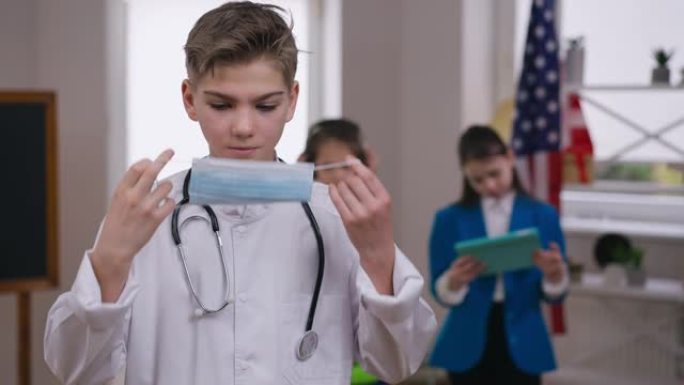 严肃的十几岁的男孩戴上冠状病毒面罩，指着拇指看着相机。自信的白人少年穿着医疗礼服的肖像，渴望在新型冠