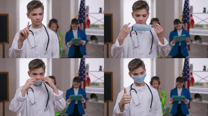 严肃的十几岁的男孩戴上冠状病毒面罩，指着拇指看着相机。自信的白人少年穿着医疗礼服的肖像，渴望在新型冠