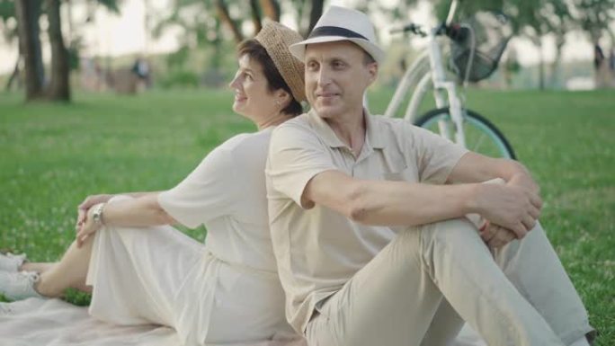 开朗的成年男女背靠背坐在绿色草坪上聊天。快乐微笑的高加索丈夫和妻子在公园户外享受夏日的肖像。爱，结合