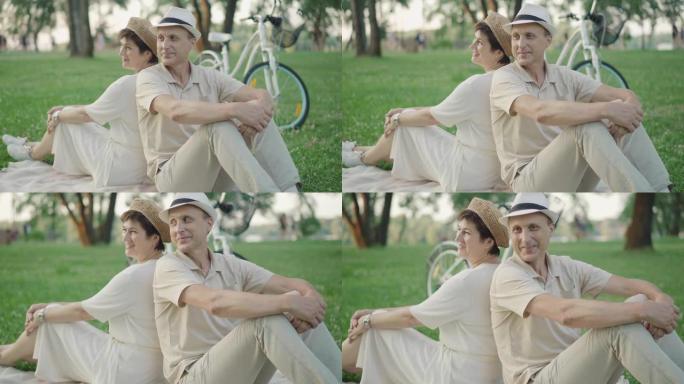 开朗的成年男女背靠背坐在绿色草坪上聊天。快乐微笑的高加索丈夫和妻子在公园户外享受夏日的肖像。爱，结合