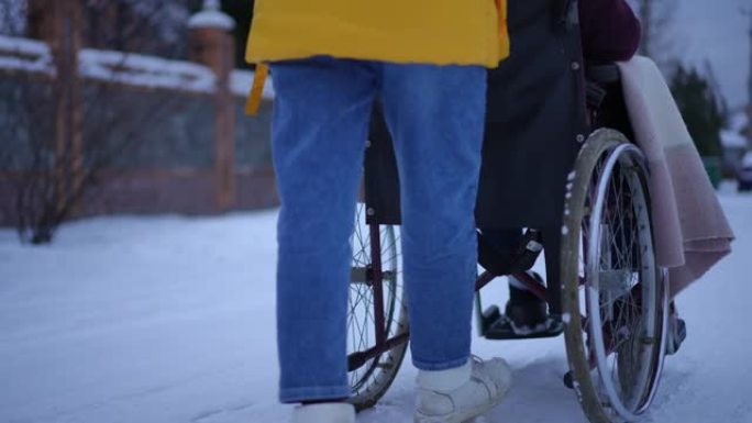 后视图无法辨认的女人在下雪的冬季道路上与男人慢动作地推着轮椅走路。年轻自信的妻子照顾丈夫在雪地上的城