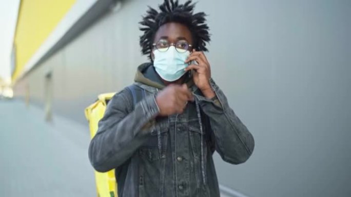 自信的非洲裔美国男子戴着新型冠状病毒肺炎的面具在电话里用绝缘的食物递送袋走路。城市专业快递订单搜索客