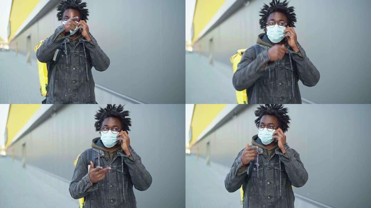 自信的非洲裔美国男子戴着新型冠状病毒肺炎的面具在电话里用绝缘的食物递送袋走路。城市专业快递订单搜索客