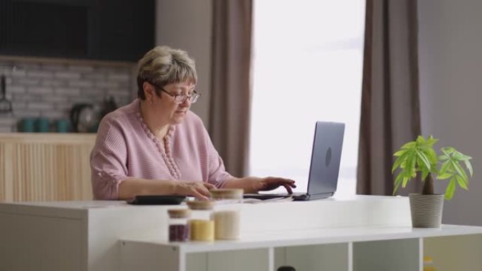 老年妇女正在家庭厨房使用笔记本电脑，检查文件并填写程序报告
