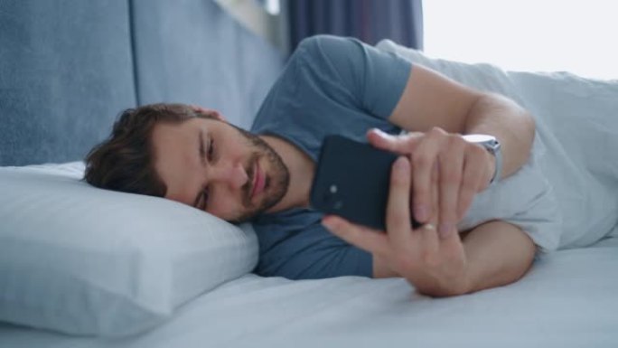 清晨躺在床上的年轻人使用智能手机。一个年轻人躺在床上，用智能手机发送信息的真实特写镜头