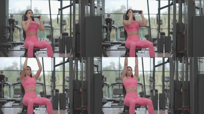 一位穿着粉红色西装的黑发西班牙裔妇女在健身房训练肩膀时，将哑铃举到顶部。坐在长凳上锻炼肩膀和手臂。重