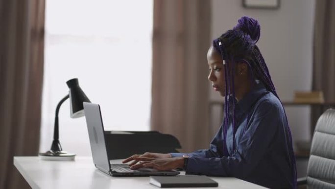 成年黑人妇女正在家里在线学习，坐在笔记本电脑的桌子旁，在键盘上打字，肖像