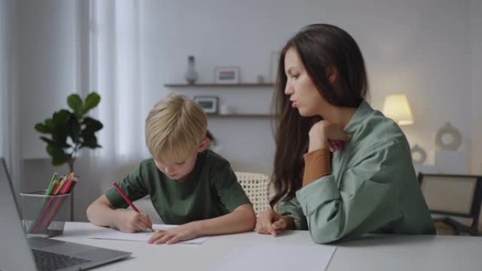 7岁的妈妈和儿子坐在客厅的桌子旁一起做作业。有爱心的年轻母亲照顾儿子的教育