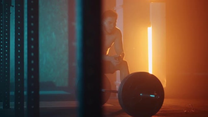 锻炼后一个疲倦的男人坐在杠铃上，在慢动作中以蓝橙色的颜色在健身房休息