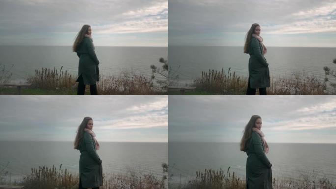 背影的混合民族妇女站在悬崖上，看着海景，转向镜头在喜怒无常的阴天。穿着绿色外套的长发女性在海边沉思。