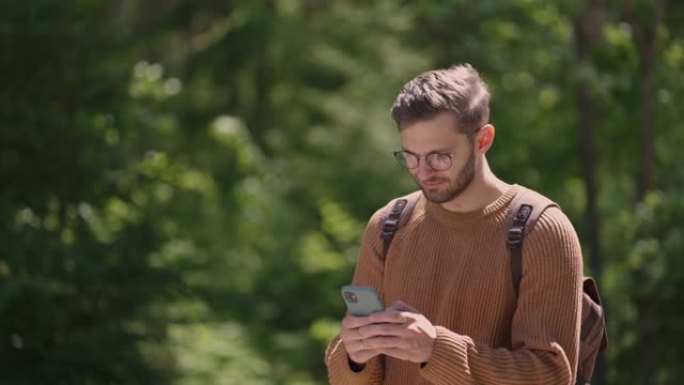 一个背着手机背包的年轻人正在森林地区散步，写信息，在线交流，观看内容，微笑着，享受散步。