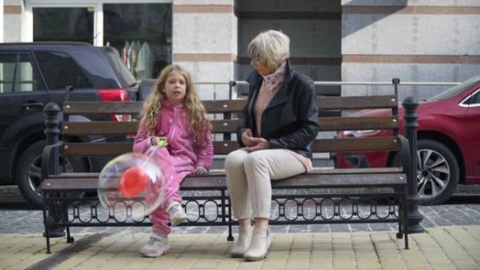 前视图美丽的奶奶和女孩坐在城市街道的长凳上聊天，女孩带着气球逃跑。宽镜头肖像轻松的高加索妇女和儿童周