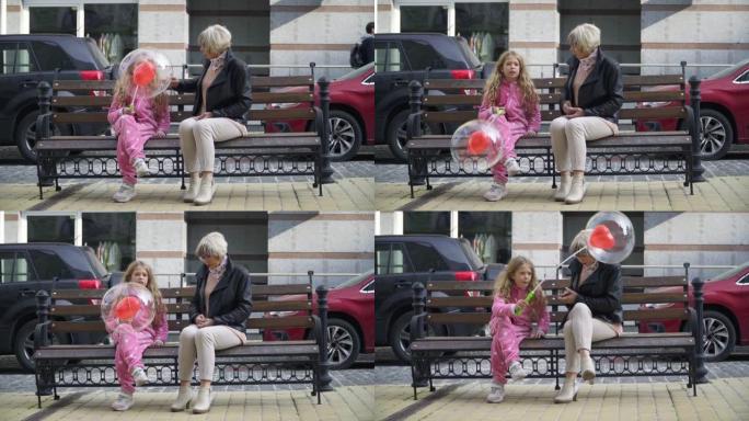 前视图美丽的奶奶和女孩坐在城市街道的长凳上聊天，女孩带着气球逃跑。宽镜头肖像轻松的高加索妇女和儿童周