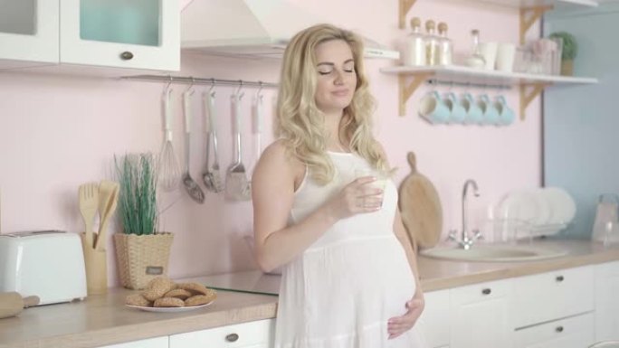 迷人的孕妇早上喝牛奶，爱抚肚子。无忧无虑快乐的高加索人孕妇怀孕期间喝健康饮料的肖像。
