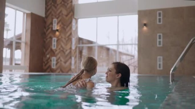 快乐的妈妈和小女孩在现代家庭水疗中心游泳池的水中游泳和玩耍
