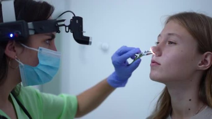 近距离侧视图阳性耳鼻喉科医师使用耳鼻喉前照灯检查患者的鼻道。专业白人妇女用鼻钳检查十几岁女孩的鼻子。