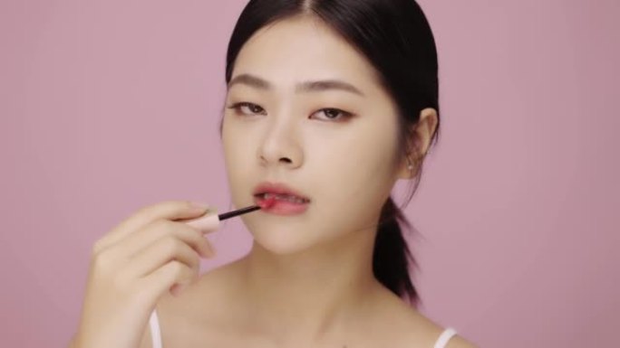 美丽的年轻亚洲女人的特写脸在粉红色背景上嘴唇上涂抹嘴唇。
