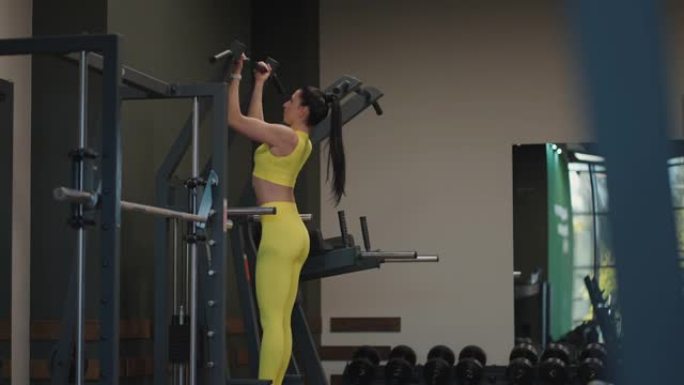 西班牙裔妇女在Gravitron上进行引体向上，以增强健身房中的肩部肌肉。一个女人正确地做练习，挺直
