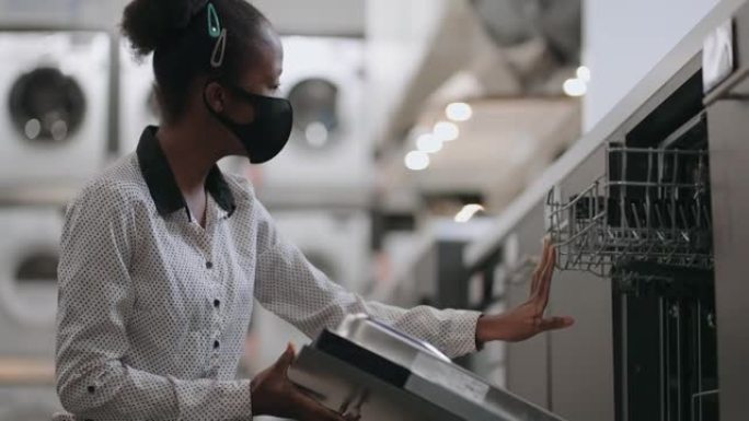 戴着口罩的年轻非洲裔美国女士正在家用电器商店购买洗碗机，查看展览样本