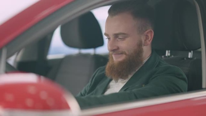 英俊的年轻人，红色的胡须和灰色的眼睛坐在新的红色汽车的驾驶员座位上，微笑着。成功的高加索商人在经销店
