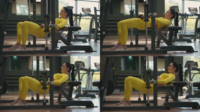一名身穿黄色运动服的年轻女子用杠铃抬起身体，训练大腿和臀部的后部。史密斯模拟器中的重量训练。在健身房