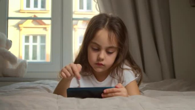 可爱的小女孩拿着智能手机在家欣赏手机电影。小孩子正在用手机学习，观看视频，享受移动技术的概念。