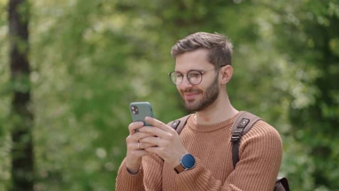 一个年轻人在树林里背着背包，在大自然中用手机写信息。在空旷地区的游客在聊天中交流，看照片，高兴，微笑