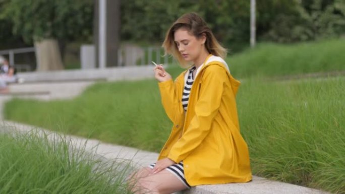 穿着黄色雨衣的美丽年轻女子享受户外吸烟在城市散步