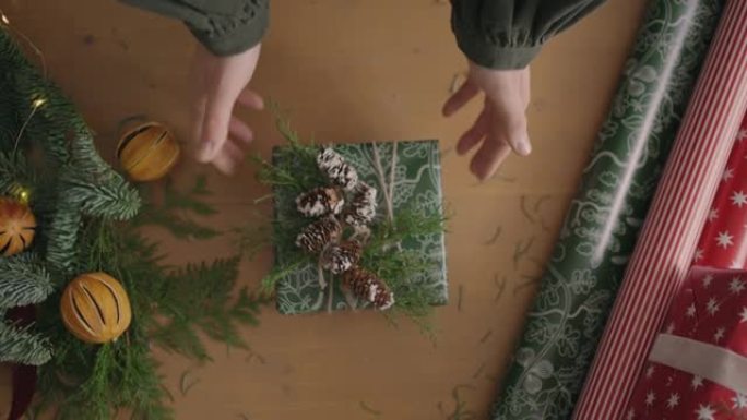 装饰者桌面的俯视图完全可见带有装饰的桌子。女性双手将用手纸包裹的圣诞礼物放在木桌上