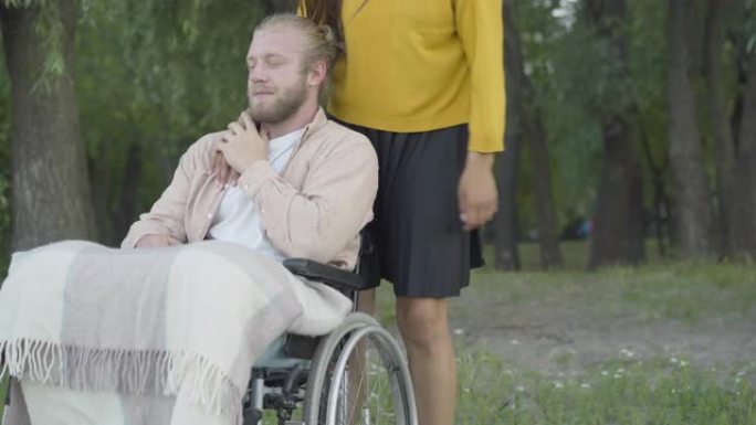 面目全非的女人来到坐在轮椅上拥抱男朋友的男人身边。晚上在夏季公园与女友约会的快乐残疾白人肖像。