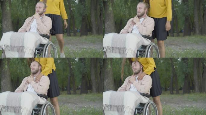 面目全非的女人来到坐在轮椅上拥抱男朋友的男人身边。晚上在夏季公园与女友约会的快乐残疾白人肖像。