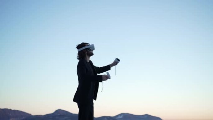 商人戴着VR虚拟现实护目镜在户外山上俯瞰日落灯光下的湖泊，环顾四周挥舞着手玩游戏