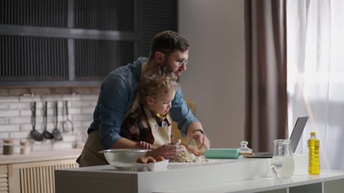 单身父亲正在学习他的小儿子做饭，用笔记本电脑在互联网上阅读食谱，在家庭厨房里煮面团