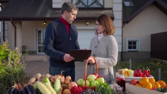 成年农民夫妇在农产品市场上争论产品清单。白人丈夫和妻子在做手势出售当地水果和蔬菜的中等镜头肖像。